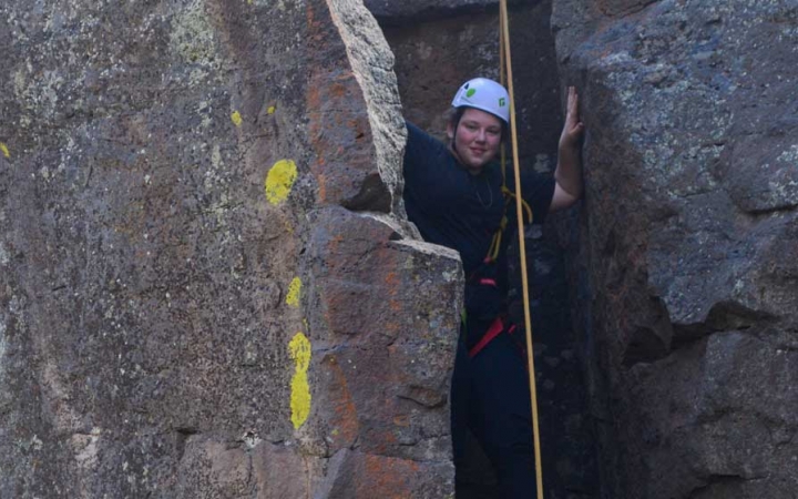A student wearing safety gear smiles from a rock crevice while rock climbing.